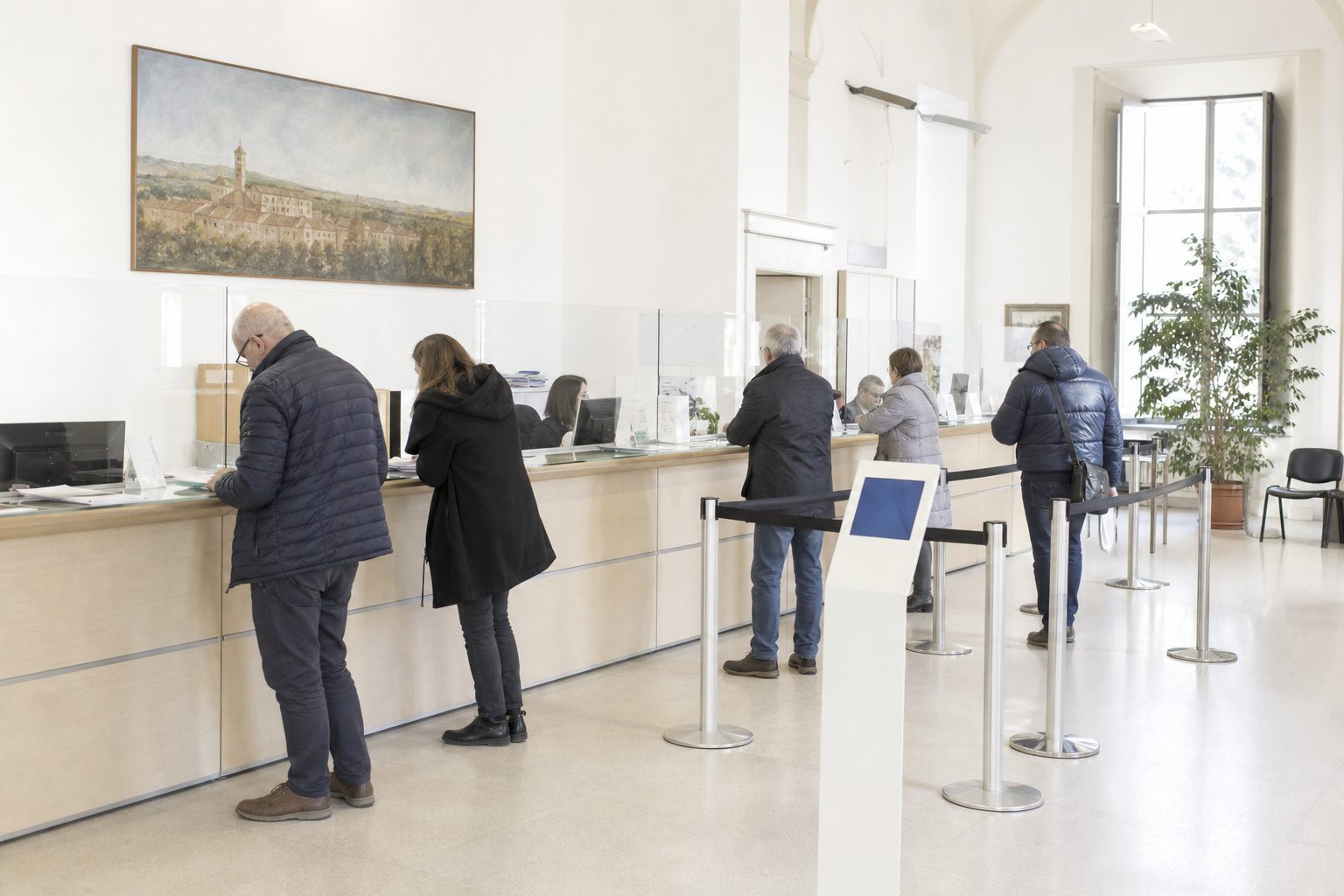 Cittadini in piedi davanti al bancone di uno sportello comunale, con le colonnine del percorso della fila e un totem per la chiamata.